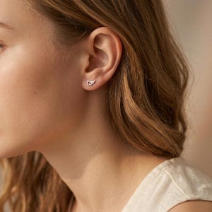 May include: A close-up of a person's ear with a silver wing-shaped stud earring. The earring is small and delicate, with a detailed wing design. The person has light skin and wavy brown hair.
