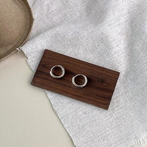 Puede incluir: Dos peque&ntilde;os pendientes de aro plateados expuestos sobre un rect&aacute;ngulo de madera oscura. Los pendientes son circulares y tienen un dise&ntilde;o simple y minimalista. El rect&aacute;ngulo de madera descansa sobre una tela blanca texturizada.