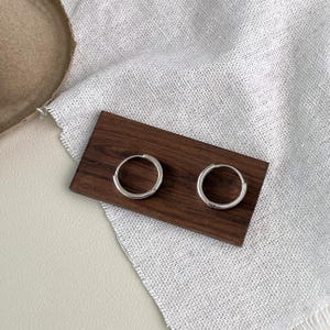 Puede incluir: Dos pendientes de aro plateados, de dise&ntilde;o circular y sencillo, presentados sobre una peque&ntilde;a tabla rectangular de madera oscura. Los pendientes se exhiben sobre una tela blanca texturizada, con un fondo neutro.