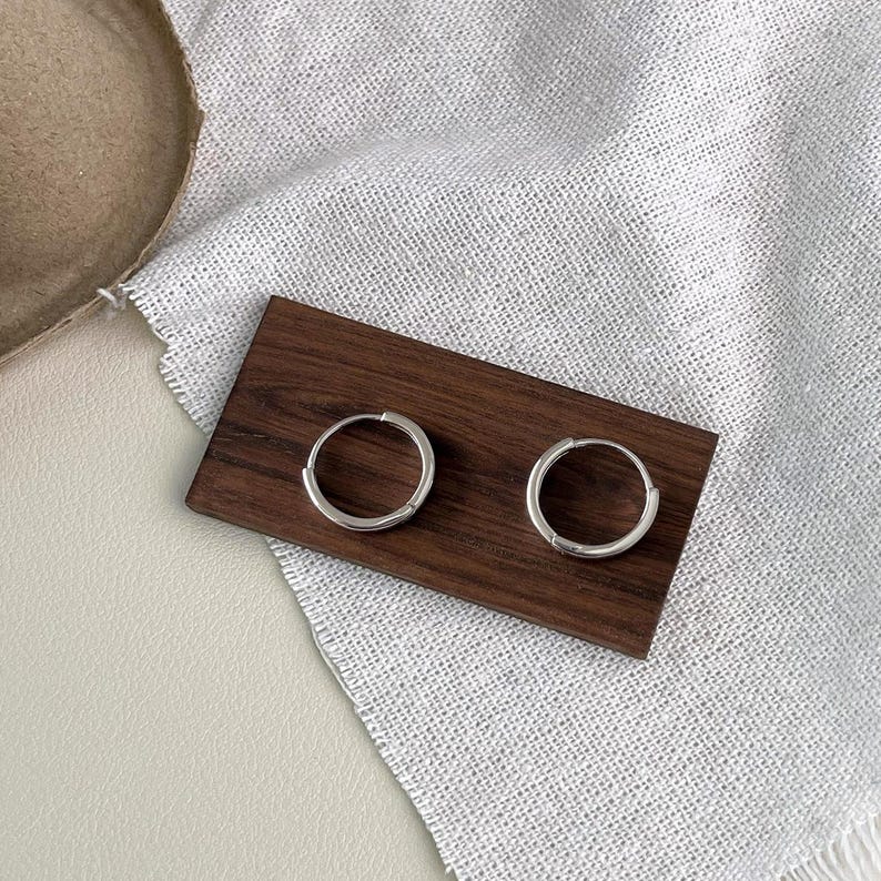 Puede incluir: Dos pendientes de aro plateados, de dise&ntilde;o circular y sencillo, presentados sobre una peque&ntilde;a tabla rectangular de madera oscura. Los pendientes se exhiben sobre una tela blanca texturizada, con un fondo neutro.