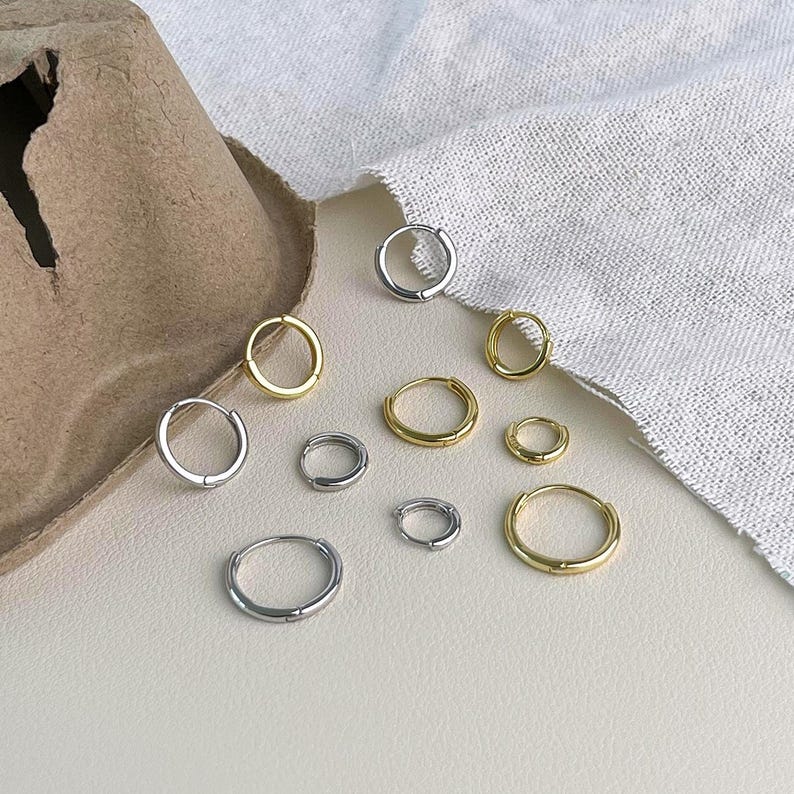 Puede incluir: Una variedad de pendientes de aro peque&ntilde;os en tonos dorados y plateados. Los pendientes var&iacute;an en tama&ntilde;o y est&aacute;n dispuestos sobre una superficie neutra con una tela blanca de fondo. Los pendientes tienen un dise&ntilde;o simple y minimalista.