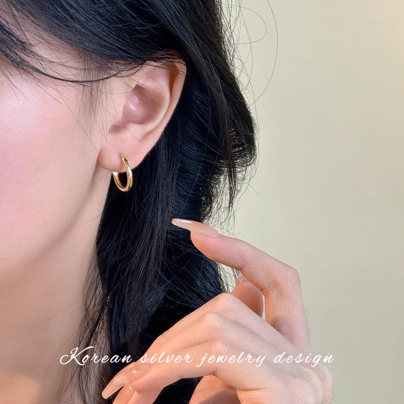 Puede incluir: Pendientes de aro dorados. Los pendientes se llevan en la oreja de una persona con cabello oscuro. El texto "Korean silver jewelry design" est&aacute; escrito en cursiva en la parte inferior de la imagen.