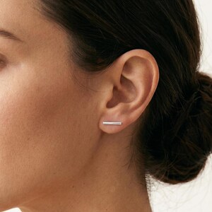Könnte beinhalten: Nahaufnahme eines Ohrs mit einem kleinen, silbernen Ohrstecker in Form eines Balkens. Der Ohrring ist ein schlichtes, minimalistisches Design, das Licht reflektiert. Der Hautton ist hell, und der Hintergrund ist unscharf.
