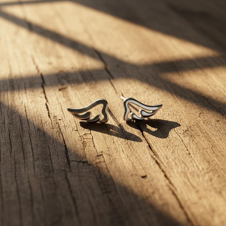 May include: A pair of silver-colored wing-shaped stud earrings. The earrings are on a wooden surface, with sunlight casting shadows across the wood. The earrings have a detailed, open design, with curved lines and a polished finish.