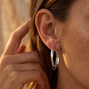May include: A pair of silver-toned hoop earrings featuring an interlocking double-loop design. The earrings are constructed from thin, polished metal bands that cross over each other to create a layered, circular silhouette worn in the earlobe.