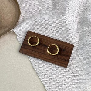 Puede incluir: Dos pendientes de aro dorados exhibidos sobre una tabla de madera marr&oacute;n oscuro. Los pendientes son circulares y tienen un dise&ntilde;o simple y minimalista. El fondo incluye una tela blanca y una superficie beige.