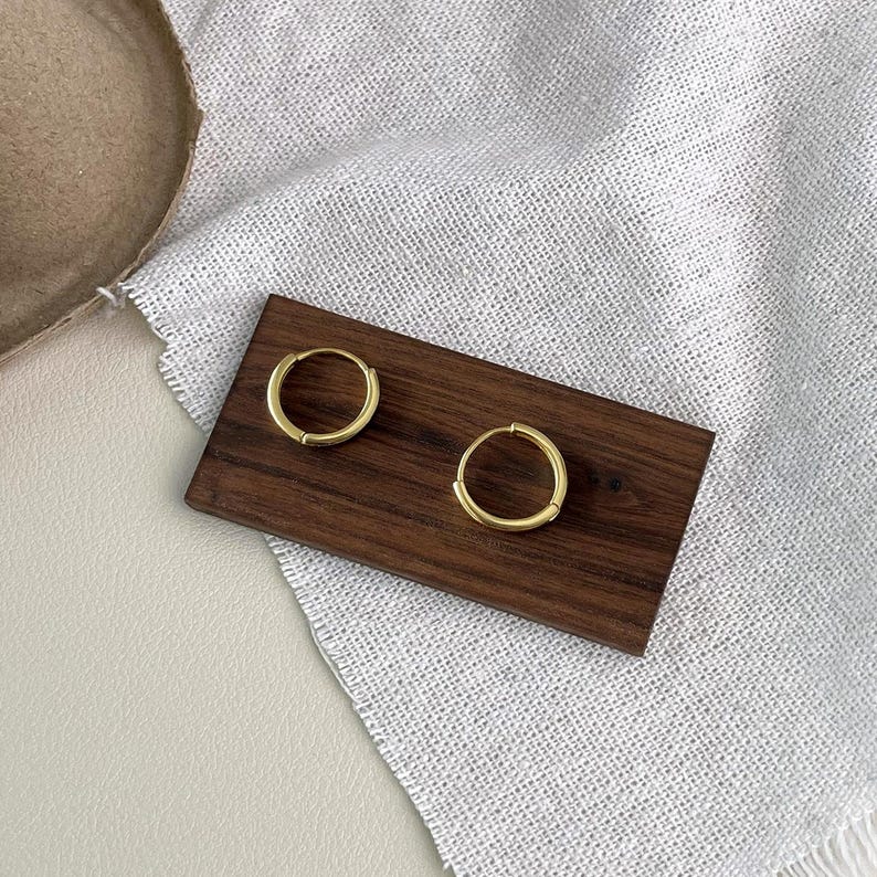Puede incluir: Dos pendientes de aro dorados exhibidos sobre una tabla de madera marr&oacute;n oscuro. Los pendientes son circulares y tienen un dise&ntilde;o simple y minimalista. El fondo incluye una tela blanca y una superficie beige.