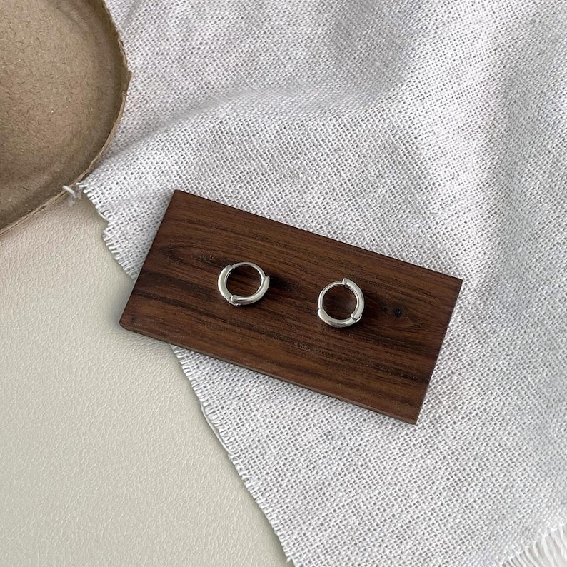 Puede incluir: Un par de peque&ntilde;os pendientes de aro plateados expuestos sobre una superficie rectangular de madera oscura. Los pendientes tienen un dise&ntilde;o simple y minimalista. La superficie de madera descansa sobre una tela blanca texturizada.