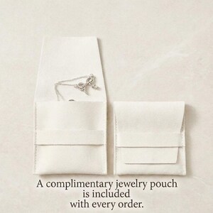 May include: Two off-white jewelry pouches, one open to reveal a silver necklace. The necklace has a bow and charm pendant. The text at the bottom reads, "A complimentary jewelry pouch is included with every order."