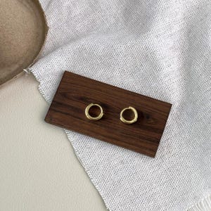 Puede incluir: Un par de pendientes de aro peque&ntilde;os, de color dorado, expuestos sobre una superficie de madera oscura. Los pendientes son circulares y tienen un dise&ntilde;o sencillo y minimalista. El fondo incluye una tela blanca texturizada y una superficie de tono neutro.