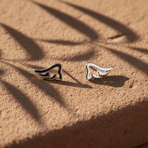 May include: A pair of silver-toned stud earrings in the shape of stylized wings. Each earring features a black outline and white enamel filling, resting on a textured, tan surface. The earrings are designed with a minimalist aesthetic.
