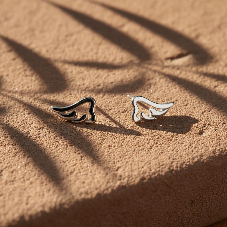 May include: A pair of silver-toned stud earrings in the shape of stylized wings. Each earring features a black outline and white enamel filling, resting on a textured, tan surface. The earrings are designed with a minimalist aesthetic.