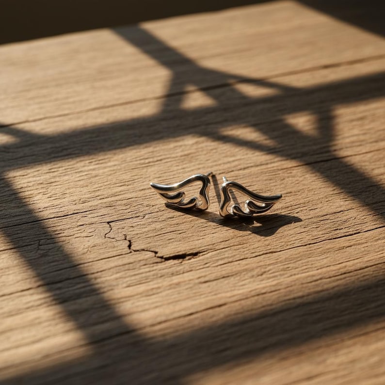 May include: A pair of silver-colored wing-shaped stud earrings. The earrings are displayed on a textured, light brown wooden surface, with shadows cast across the wood. The earrings are small and detailed, with a polished finish.