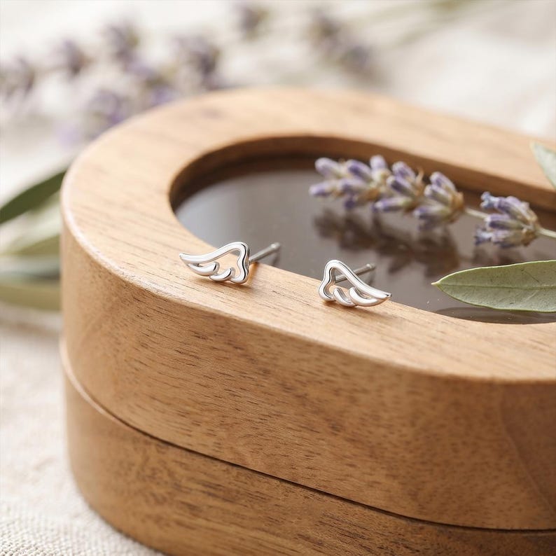 May include: A pair of small, silver-colored wing-shaped stud earrings. The earrings are displayed on a wooden box with a dark, reflective surface. Lavender sprigs and a green leaf are in the background.