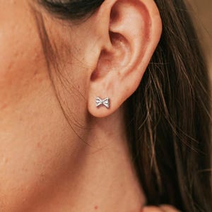May include: A close-up shot of a silver bow-shaped stud earring. The earring is small and delicate, with a detailed bow design. The earring is set against the backdrop of a person's ear and skin.