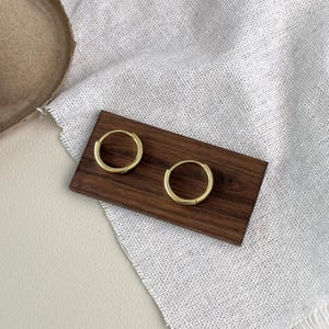 Puede incluir: Dos pendientes de aro dorados se muestran sobre un peque&ntilde;o rect&aacute;ngulo de madera oscura. Los pendientes son circulares y tienen un dise&ntilde;o simple y minimalista. El rect&aacute;ngulo de madera descansa sobre una tela texturizada blanca y gris.