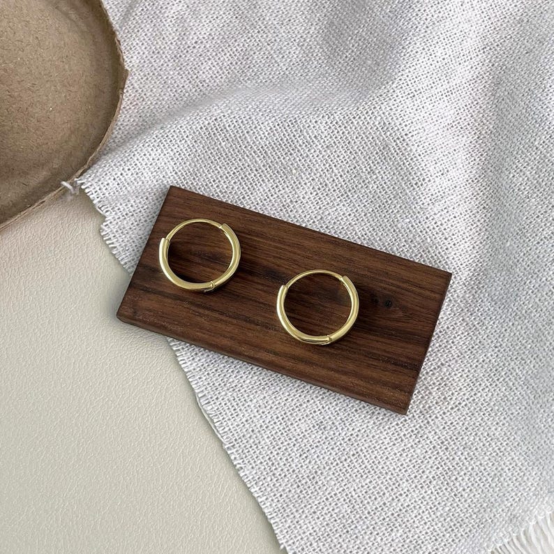 Puede incluir: Dos pendientes de aro dorados se muestran sobre un peque&ntilde;o rect&aacute;ngulo de madera oscura. Los pendientes son circulares y tienen un dise&ntilde;o simple y minimalista. El rect&aacute;ngulo de madera descansa sobre una tela texturizada blanca y gris.