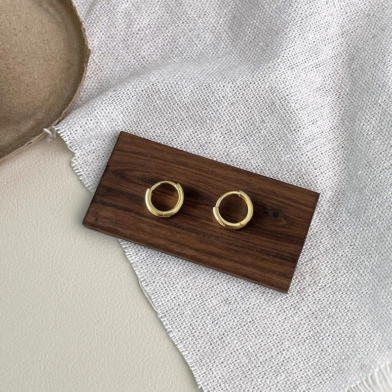 Puede incluir: Dos peque&ntilde;os pendientes de aro dorados expuestos sobre una superficie rectangular de madera oscura. Los pendientes son circulares y tienen un dise&ntilde;o simple y minimalista. La superficie de madera descansa sobre una tela blanca.