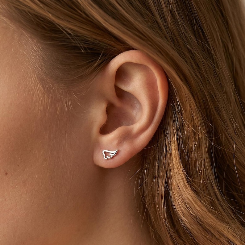 May include: A close-up of a person's ear with a pair of small, silver wing-shaped stud earrings. The earrings are simple in design, with a delicate outline of a wing. The earrings are approximately 0.3 inches in length.