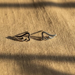 May include: A pair of silver-colored wing-shaped stud earrings. Each earring features a detailed wing design with a dark, contrasting outline. The earrings are displayed on a textured, tan-colored surface.