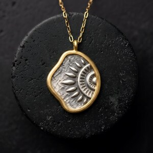 Può includere: Una collana con pendente color oro caratterizzata da una cornice di forma organica irregolare. Il centro presenta una superficie argentata e testurizzata con un motivo in rilievo simile a un sole, composto da gocce radianti e cerchi concentrici.