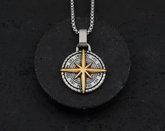 May include: Silver-tone compass pendant necklace with a gold-colored starburst design. The pendant has a detailed outer ring with the number "101" repeated. The necklace is displayed on a dark, textured surface.
