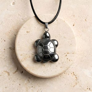 May include: A silver-coloured turtle pendant necklace with a black cord. The turtle pendant has a detailed shell design and rounded limbs. The necklace is displayed on a beige, circular stone.