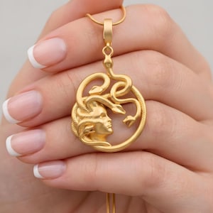 May include: Gold-colored pendant necklace featuring a Medusa design. The pendant depicts Medusa's profile surrounded by coiling snakes within a circular frame. The necklace has a delicate chain.