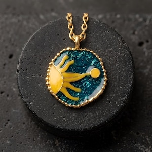 May include: A gold-colored necklace with a pendant featuring a yellow sun design on a teal background. The sun has stylized rays and a small yellow circle. The pendant is round with a gold-colored border and hangs from a gold chain.