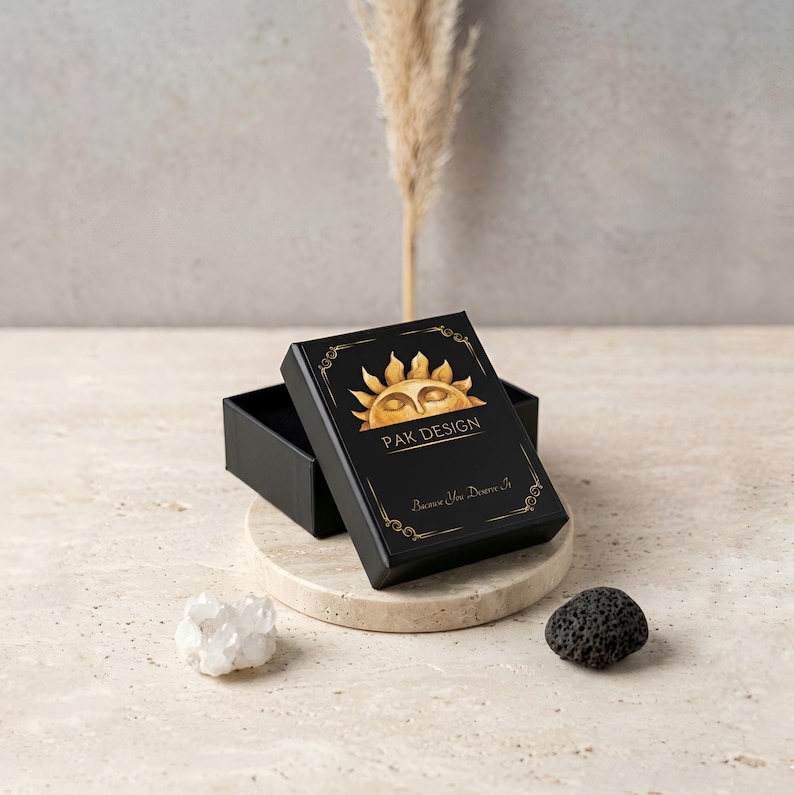 Elegant black jewelry gift box by Pak Design Jewelry featuring a golden sun logo and the text Because You Deserve It. The box is displayed on a natural stone pedestal with aesthetic crystals, ready for gifting a unique necklace to a loved one. Premium packaging for special occasion gifts.