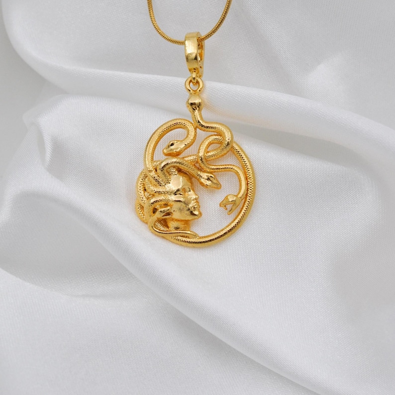 Puede incluir: Un collar con colgante dorado con un dise&ntilde;o de Medusa. El colgante muestra el perfil de Medusa con serpientes, suspendido de una fina cadena dorada. El collar se presenta sobre tela blanca y sedosa.