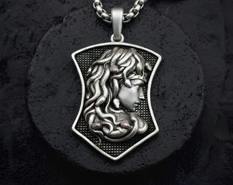 May include: Silver-toned pendant necklace featuring a detailed profile of a woman with flowing hair. The pendant is shield-shaped with a textured background and a silver chain. The necklace rests on a dark, textured surface.