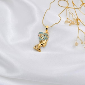 Op de afbeelding: Gouden hangerketting met een gedetailleerde buste van Nefertiti. De hanger heeft een turquoise en gouden hoofdtooi en hangt aan een fijne gouden ketting. De ketting wordt getoond op een witte satijnen achtergrond.