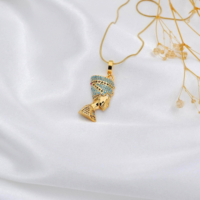Op de afbeelding: Gouden hangerketting met een gedetailleerde buste van Nefertiti. De hanger heeft een turquoise en gouden hoofdtooi en hangt aan een fijne gouden ketting. De ketting wordt getoond op een witte satijnen achtergrond.