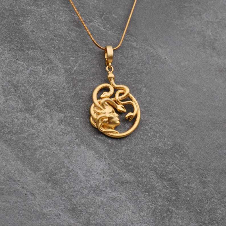 May include: Gold-colored pendant necklace featuring a Medusa design. The pendant depicts a woman's profile surrounded by snakes. The necklace has a delicate chain and a simple clasp. The jewelry is set against a gray background.
