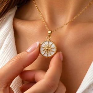 Op de afbeelding: Een goudkleurige ketting met een ronde hanger voorzien van een witte parelmoeren inleg. De hanger toont een verhoogd gouden zonmotief met uitstralende stralen en een centrale bol, hangend aan een fijne goudkleurige schakelketting.