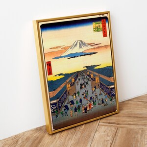 May include: A framed woodblock print of a Japanese landscape with Mount Fuji in the background. The print depicts a bustling street scene with people walking and shops lining the street. The print is in a gold frame.