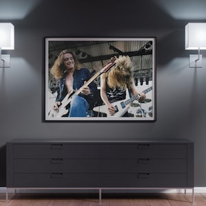 Może przedstawiać: Czarno-białe zdjęcie dwóch osób grających na gitarach elektrycznych na scenie. Zdjęcie wisi na ścianie nad ciemnoszarą komodą z sześcioma szufladami.