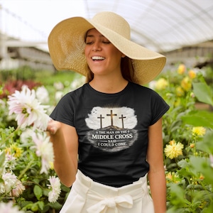 May include: Black t-shirt featuring a graphic of three crosses on a hill with the text "THE MAN ON THE MIDDLE CROSS SAID - I COULD COME." The shirt is worn by a person wearing a straw hat and white skirt, surrounded by flowers.
