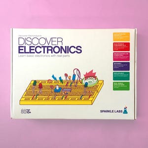 May include: A white box with the words "DISCOVER ELECTRONICS" and "Learn basic electronics with real parts." A yellow breadboard with various electronic components is illustrated. The box also features colorful labels and the Sparkle Labs logo.