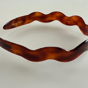 May include: A tortoiseshell-patterned headband with a wavy design. The headband is a warm brown color with darker brown accents. The text "Charles Webb, Hand Made in France" is printed on the headband.