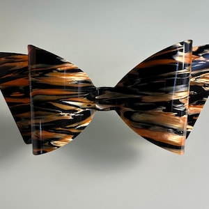 May include: A large, decorative bow with a glossy finish. The bow features a striking pattern of black, orange, and gold swirls, creating a marbled effect. The bow is a statement piece, perfect for hair accessories or gift wrapping.