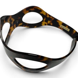 May include: A curved, tortoiseshell-patterned headband featuring large, oval-shaped cutouts throughout its structure. The inner surface displays a small gold-colored label with the text "HAND MADE IN FRANCE" and a small logo mark.