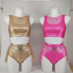 Peut inclure: Deux costumes de danse sur mannequins, un doré et l'autre rose. Chaque ensemble comprend un crop top et un short taille haute avec une superposition de jupe transparente. Les hauts et les shorts sont ornés de strass. Le texte dit : "Ces options de couleurs ont été générées par l'IA."