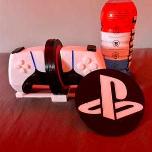 May include: A white and black video game controller with blue accents, a black coaster with a white PlayStation logo, and a red, white, and blue beverage bottle. The controller has a black ring around the middle.