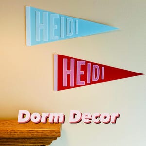 May include: Two pennant-shaped wall hangings, one light blue and one red, each with the name "HEIDI" in white letters. Below, the words "Dorm Decor" are displayed in pink letters, suggesting a theme of college or dorm room decoration.
