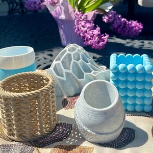May include: A collection of decorative planters in various shapes and colors. Includes a woven basket-style planter, a blue bubble-textured cube, a white and blue layered cylinder, a light blue mountain-shaped planter, and a textured gray vase. A bouquet of purple flowers is in the background.