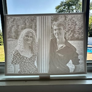 May include: A backlit, rectangular photo frame displaying a black and white image of two people. The woman on the left has curly hair and is wearing a floral top. The man on the right is wearing a button-down shirt. A pillar is between them.