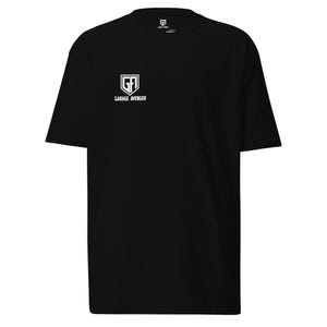 May include: Black t-shirt with a white logo on the front that says "GARAGE AVENGER". The shirt has a crew neck and short sleeves. The logo is a shield design with the letters "GF" inside.