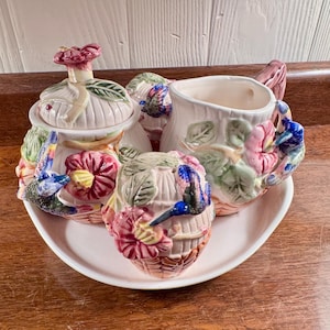 Op de afbeelding: Een keramische theeset met een kolibrie- en bloemenontwerp. De set bevat een melkkan, een suikerpot en twee andere containers, allemaal op een witte ovale schaal. De stukken zijn versierd met roze, blauwe en groene accenten.